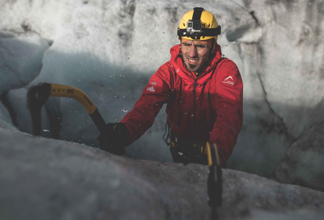 A man wearing a yellow helmet climbing an ice wall using rops and an iceax